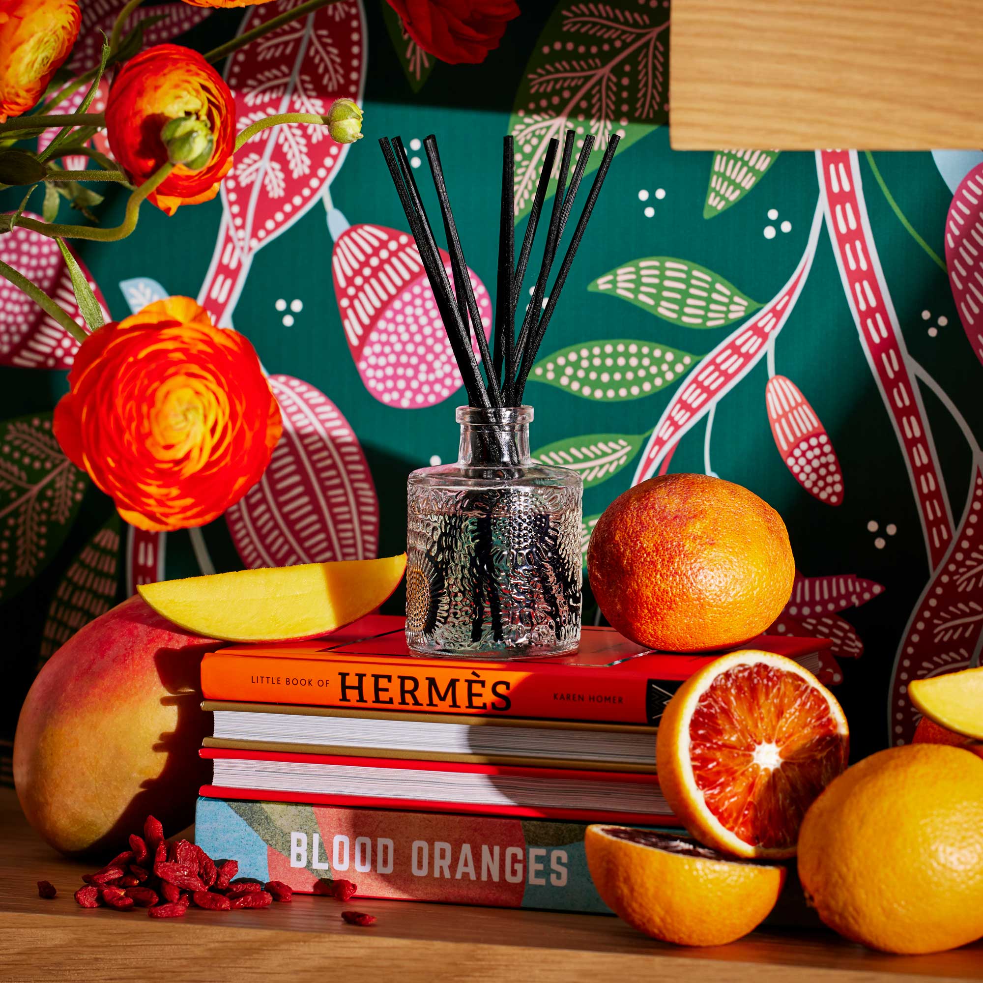 Reed diffuser on a stack of books with blood oranges and flowers against a colorful patterned background